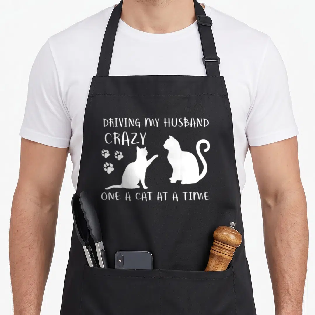 Husband cat driving aprons - White text reading DRIVING MY HUSBAND CRAZY with two cat silhouettes and three paw prints — shown on a black cotton canvas apron against a white background