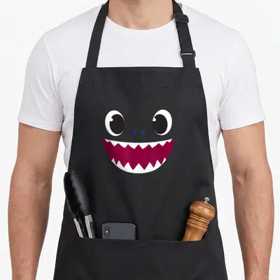 Baby daddy apron - Cartoon shark face with two eyes and jagged maroon mouth on black fabric — shown on a black cotton canvas apron against a white background