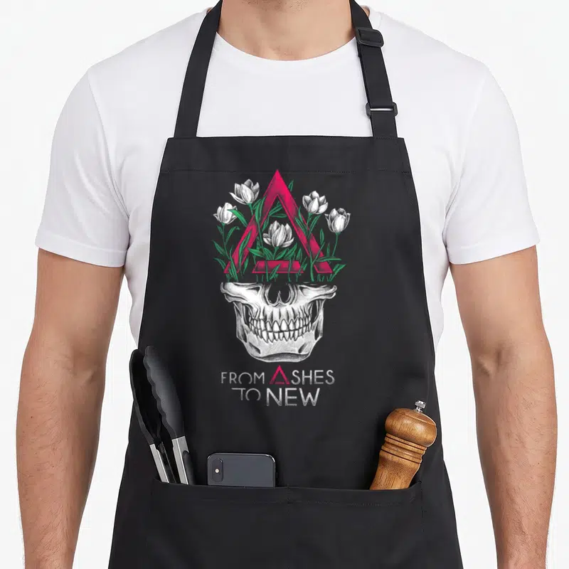 Ash face apron - White skull with pink triangle and green leaves, text FROM ASHES TO NEW — shown on a black cotton canvas apron against a white background