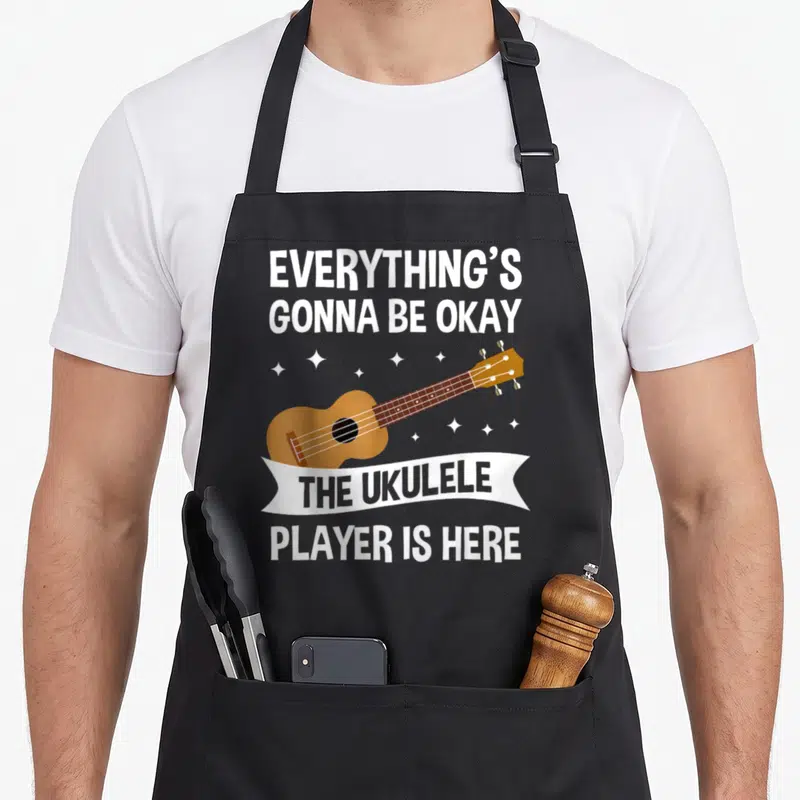 Player ukulele apron - Cartoon brown ukulele with white stars and grey text reading everything's gonna be okay — shown on a black cotton canvas apron against a white background