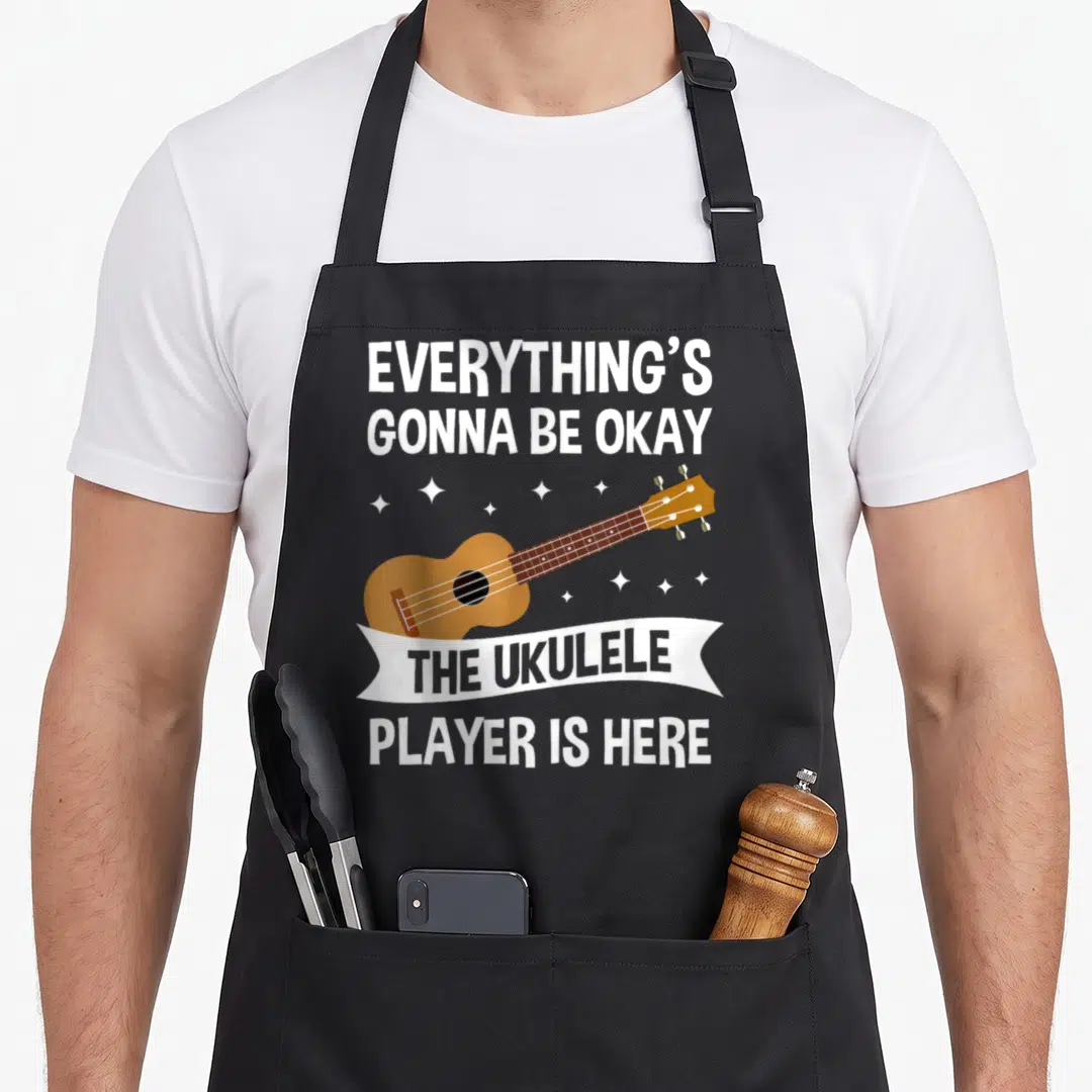 Player ukulele apron - Cartoon brown ukulele with white stars and grey text reading everything's gonna be okay — shown on a black cotton canvas apron against a white background