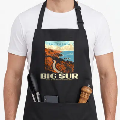 Sur bike life apron - Retro Big Sur coastline illustration with California text in orange blue and cream — shown on a black cotton canvas apron against a white background