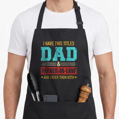 Title law father day apron - I have two titles dad father-in-law text stacked in yellow blue red distressed font — shown on a black cotton canvas apron against a white background