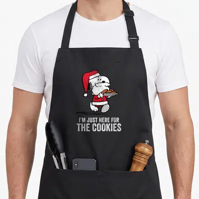Funny cooky women's christmas aprons - Snoopy Santa holding cookie plate with I'M JUST HERE FOR THE COOKIES text — shown on a black cotton canvas apron against a white background