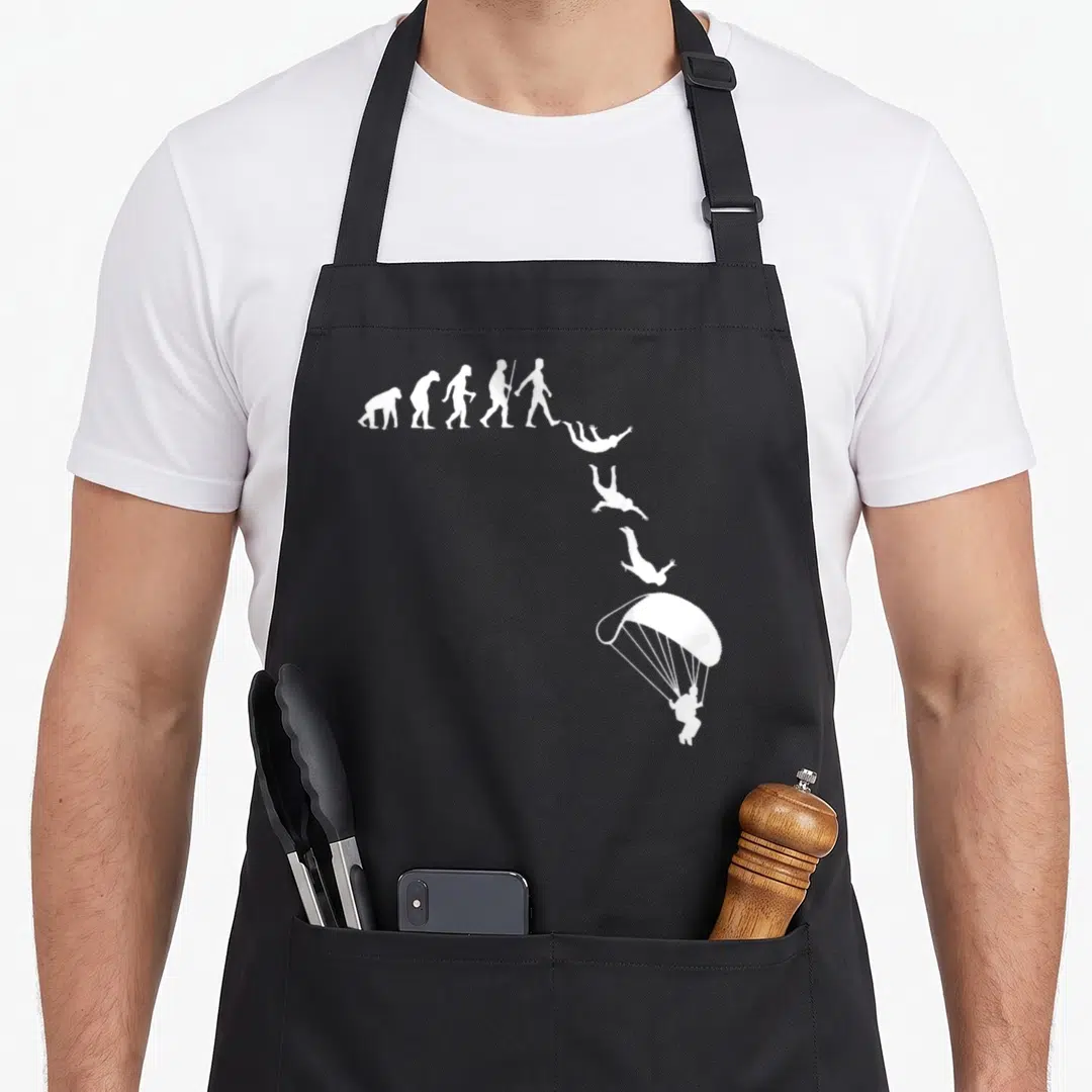 Skydive paragliding apron - White evolution silhouettes curving into a falling parachutist on black fabric — shown on a black cotton canvas apron against a white background