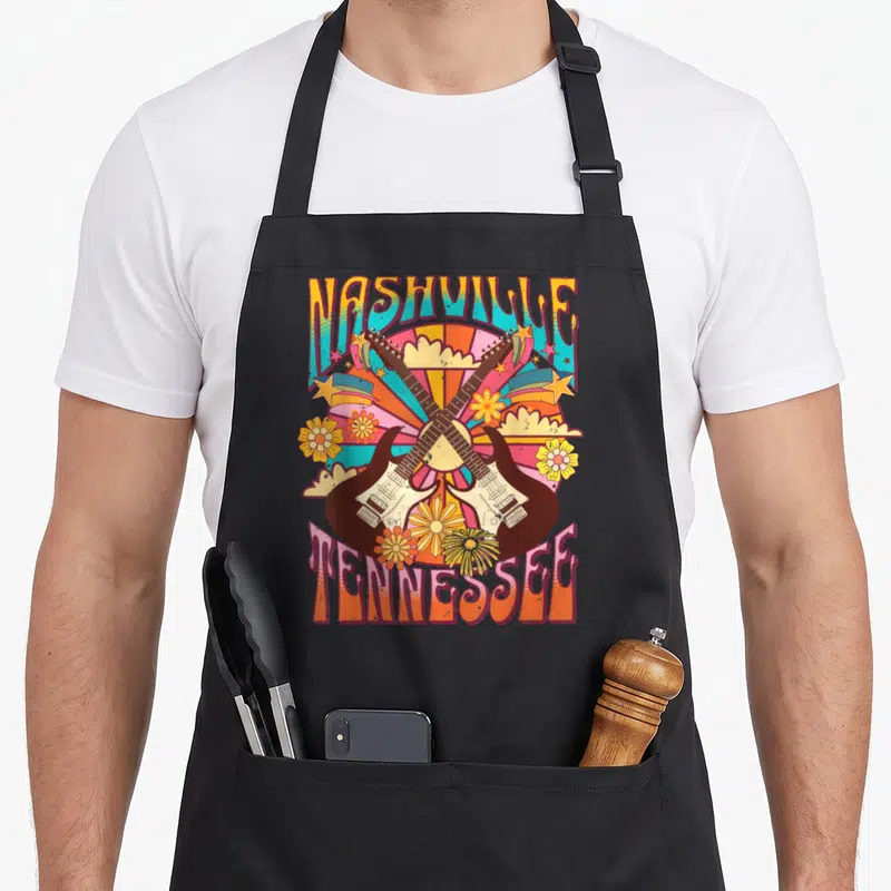 Nash country apron - Crossed electric guitars with Nashville Tennessee text and retro flowers in orange and teal — shown on a black cotton canvas apron against a white background