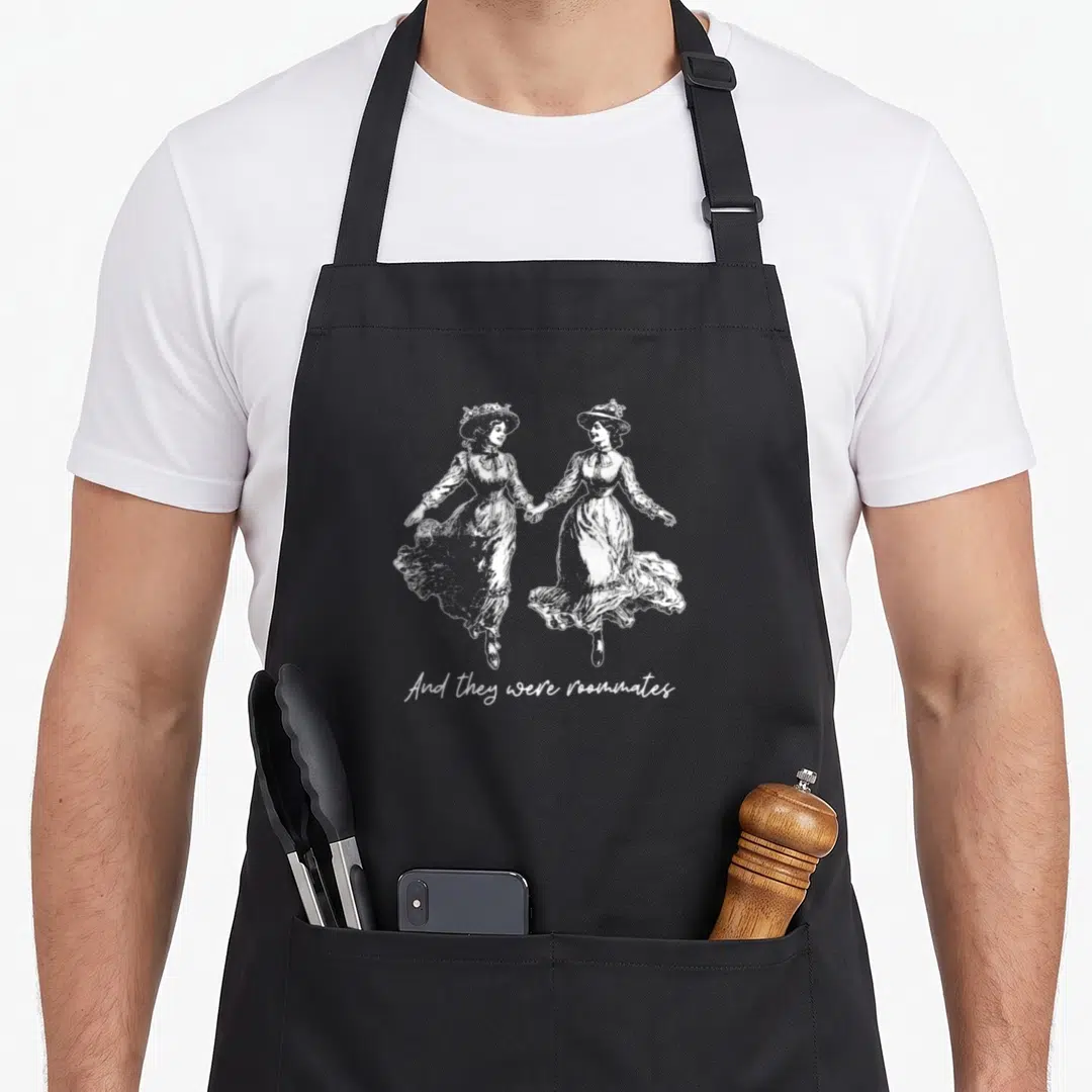 They pride apron - White line art of two women in vintage dresses holding hands above text reading And they were — shown on a black cotton canvas apron against a white background