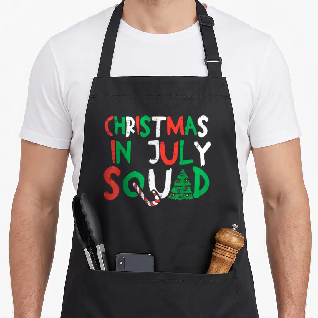 July christmas apron - Distressed red green white text reading Christmas In July Squad with candy cane and tree — shown on a black cotton canvas apron against a white background