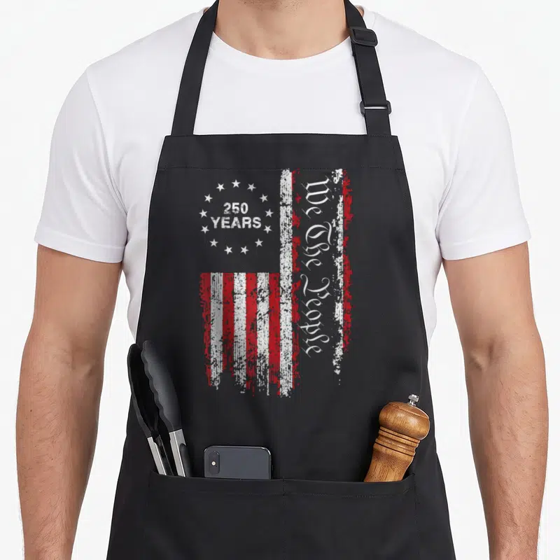 250th independence apron - Distressed USA flag with 250 YEARS in stars and vertical We The People text in red and white — shown on a black cotton canvas apron against a white background