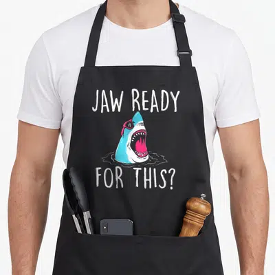 Jaw ocean apron - Cartoon shark head in blue and pink with JAW READY FOR THIS? text — shown on a black cotton canvas apron against a white background