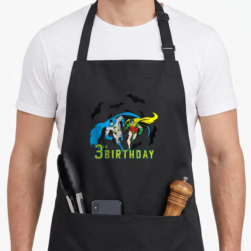 Robin birthday apron - Batman and Robin running with black bats and 3rd BIRTHDAY text in blue yellow red and black — shown on a black cotton canvas apron against a white background