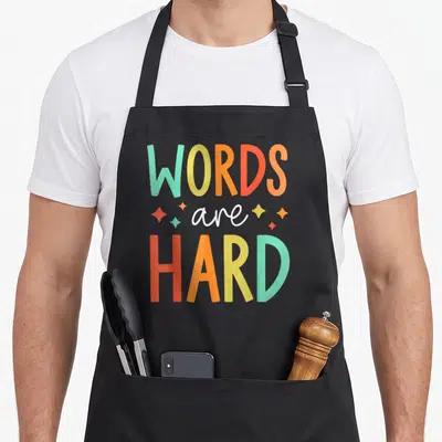 Pathology teacher apron - Colorful WORDS are HARD text with scattered stars in teal, yellow, orange, and red — shown on a black cotton canvas apron against a white background