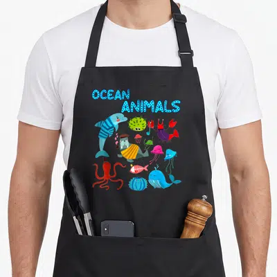 Creature animal apron - Cartoon ocean animals including dolphin, octopus, and walrus under blue OCEAN ANIMALS text — shown on a black cotton canvas apron against a white background