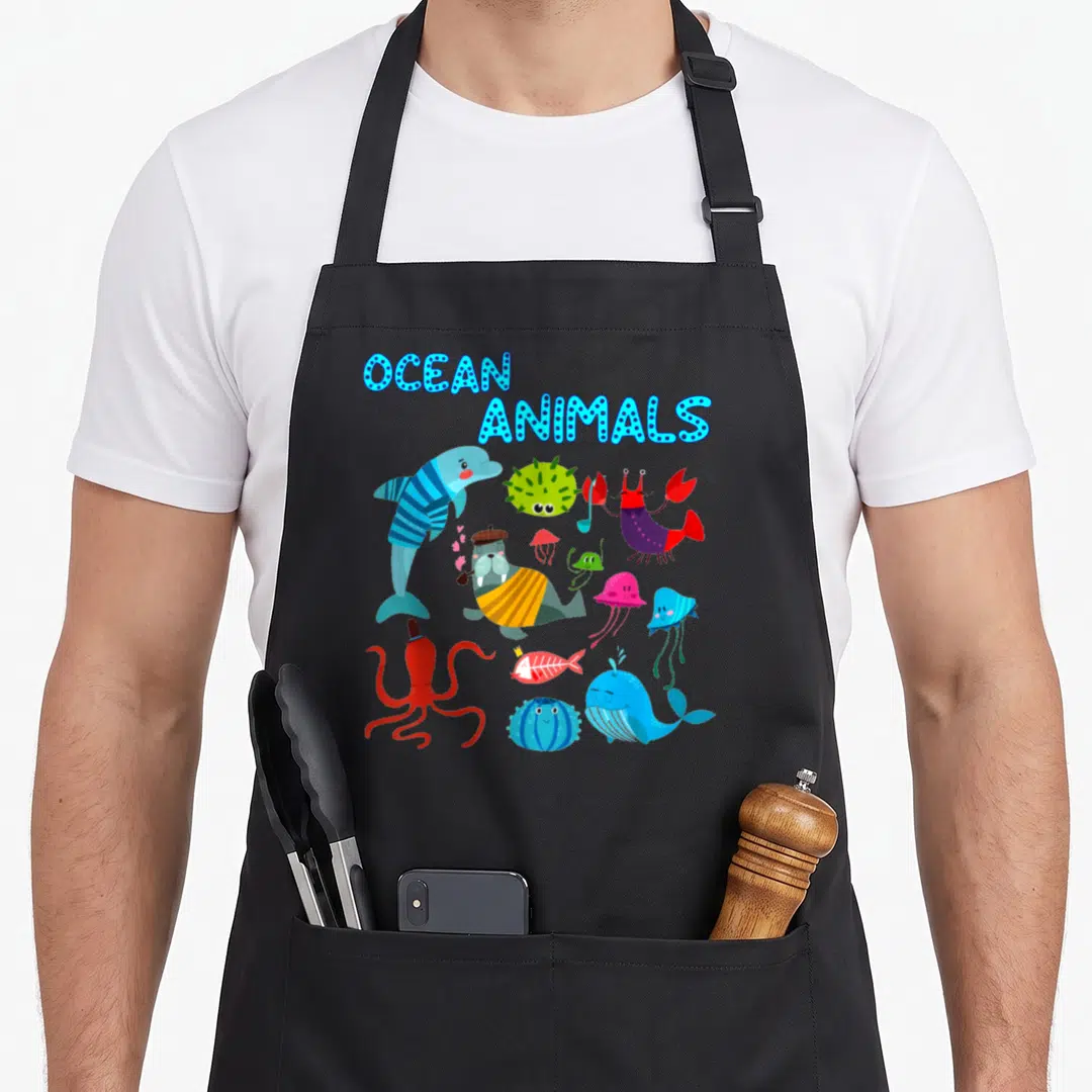 Creature animal apron - Cartoon ocean animals including dolphin, octopus, and walrus under blue OCEAN ANIMALS text — shown on a black cotton canvas apron against a white background