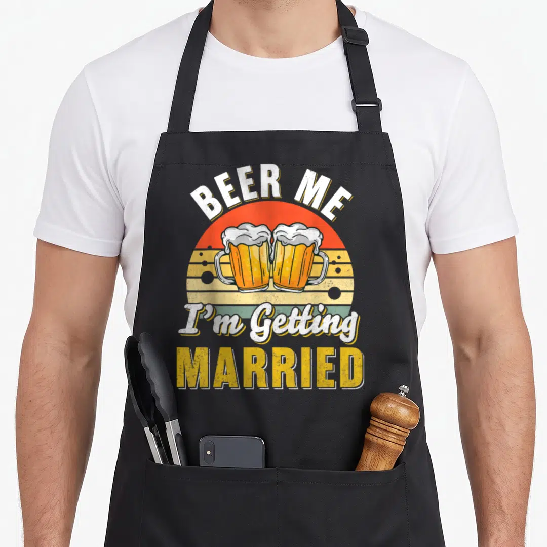 Supply funny wedding aprons - Two clinking beer mugs with BEER ME I'm Getting MARRIED text over retro sun — shown on a black cotton canvas apron against a white background