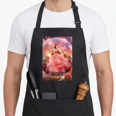 Glinda wicked apron - Woman in pink dress on gold throne with wand, bubbles, castle, and WICKED text — shown on a black cotton canvas apron against a white background