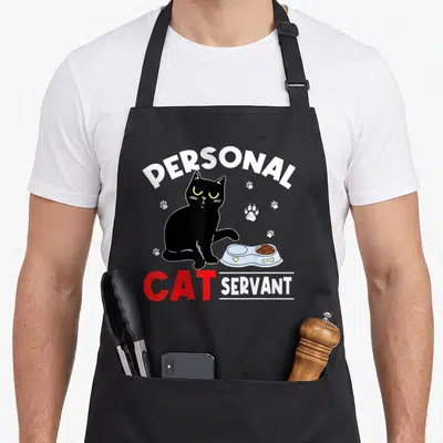 Servant personal printed aprons - Cartoon black cat sits by blue food bowl with PERSONAL CAT SERVANT text in red — shown on a black cotton canvas apron against a white background
