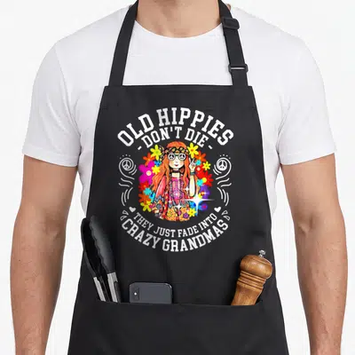 Hippy funny grandma apron - Cartoon hippie woman with flowers and peace signs surrounded by Old Hippies Don't Die text — shown on a black cotton canvas apron against a white background