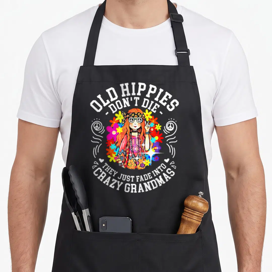 Hippy funny grandma apron - Cartoon hippie woman with flowers and peace signs surrounded by Old Hippies Don't Die text — shown on a black cotton canvas apron against a white background