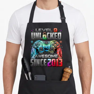9th gaming aprons - Level 9 Unlocked Awesome Since 2013 text with rainbow game controller graphic — shown on a black cotton canvas apron against a white background