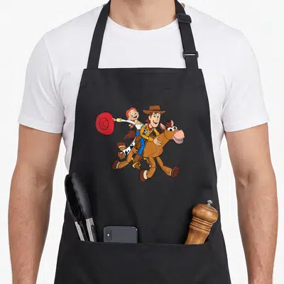 Roundup dog breed aprons - Cartoon of Woody and Jessie riding Bullseye galloping right holding red hat — shown on a black cotton canvas apron against a white background