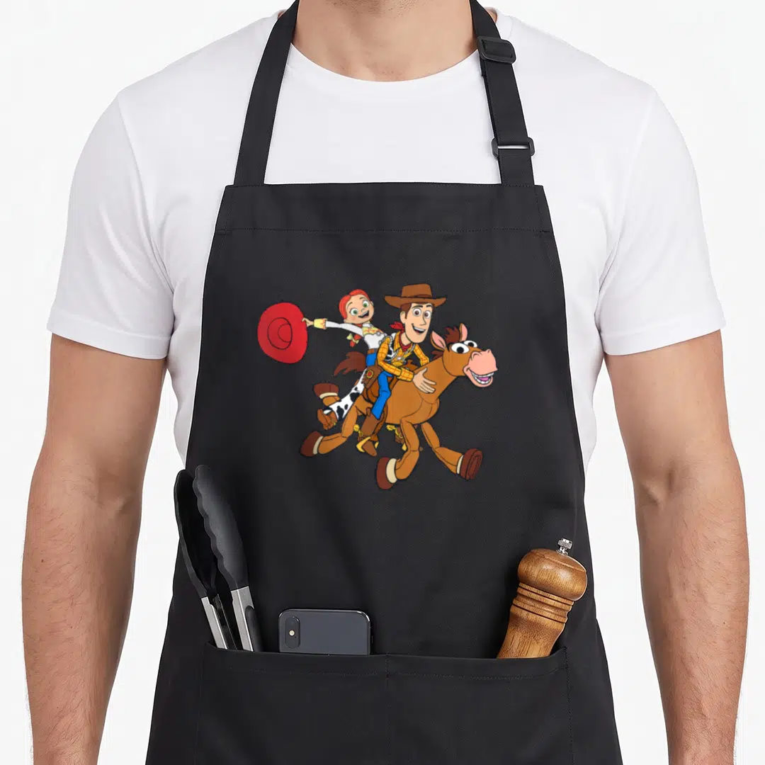 Roundup dog breed aprons - Cartoon of Woody and Jessie riding Bullseye galloping right holding red hat — shown on a black cotton canvas apron against a white background