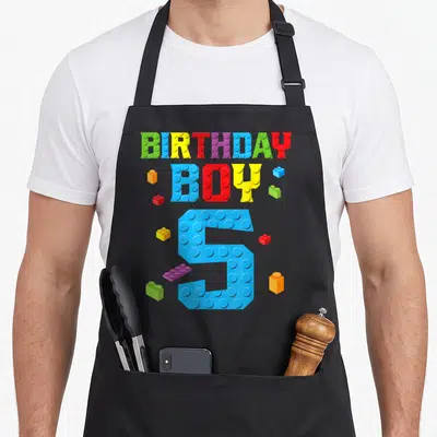 5th grill master apron - BIRTHDAY BOY 5 text in brick style with scattered toy bricks in blue red yellow green purple — shown on a black cotton canvas apron against a white background
