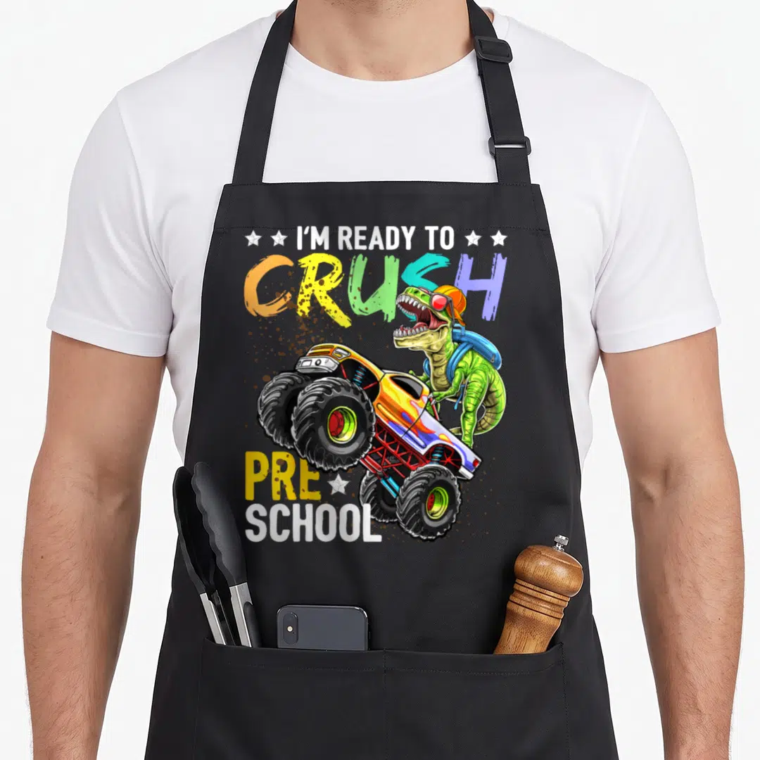 Dinosaur truck apron - Green dinosaur on monster truck with I'M READY TO CRUSH PRE SCHOOL text in multi-color letters — shown on a black cotton canvas apron against a white background