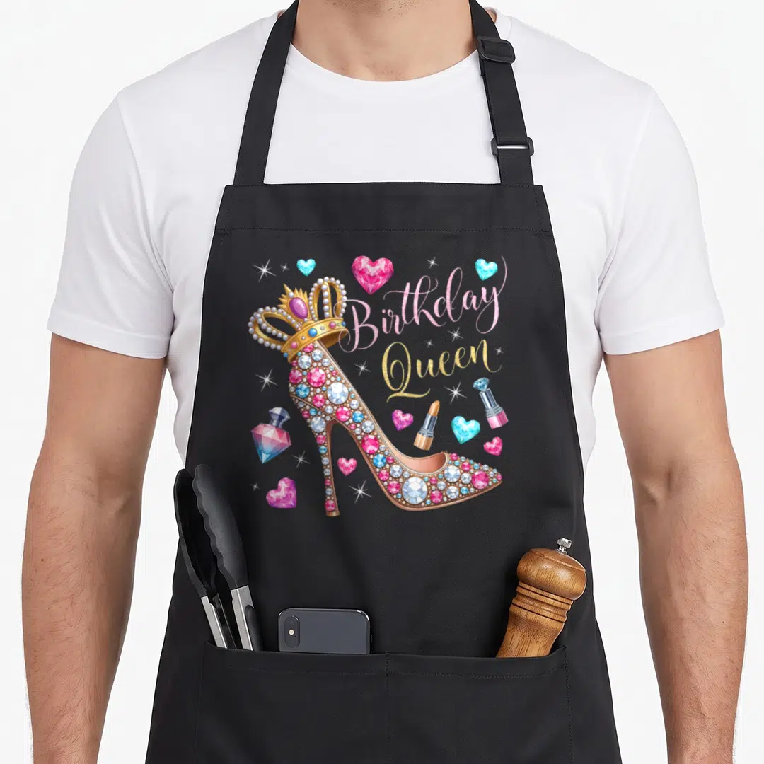 Heel queen of baking apron - Jeweled high heel shoe with crown and birthday queen text surrounded by gems and cosmetics — shown on a black cotton canvas apron against a white background