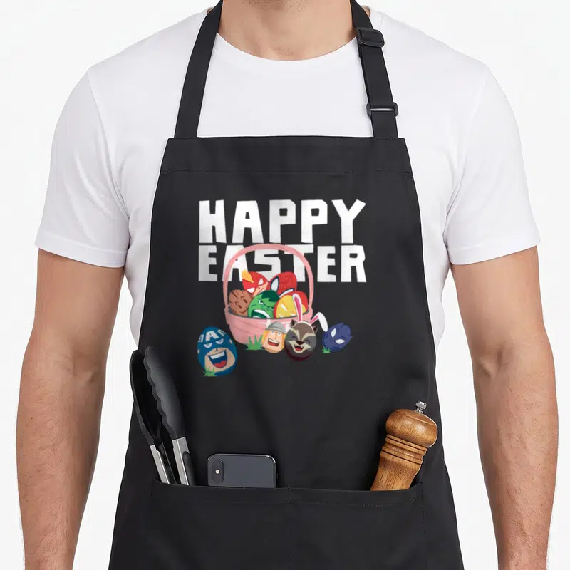 Funny egg basket apron - HAPPY EASTER text above pink basket with character eggs and grass patches — shown on a black cotton canvas apron against a white background