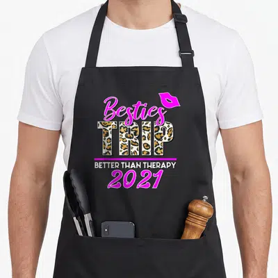 Trip leopard apron - Besties TRIP BETTER THAN THERAPY 2021 text with leopard print fill and pink lips — shown on a black cotton canvas apron against a white background