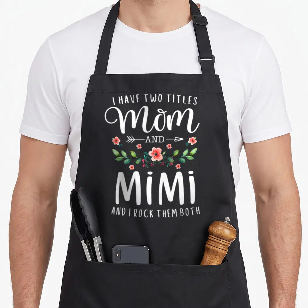 Title floral apron - White text I Have Two Titles Mom and Mimi with pink flowers and green leaves — shown on a black cotton canvas apron against a white background