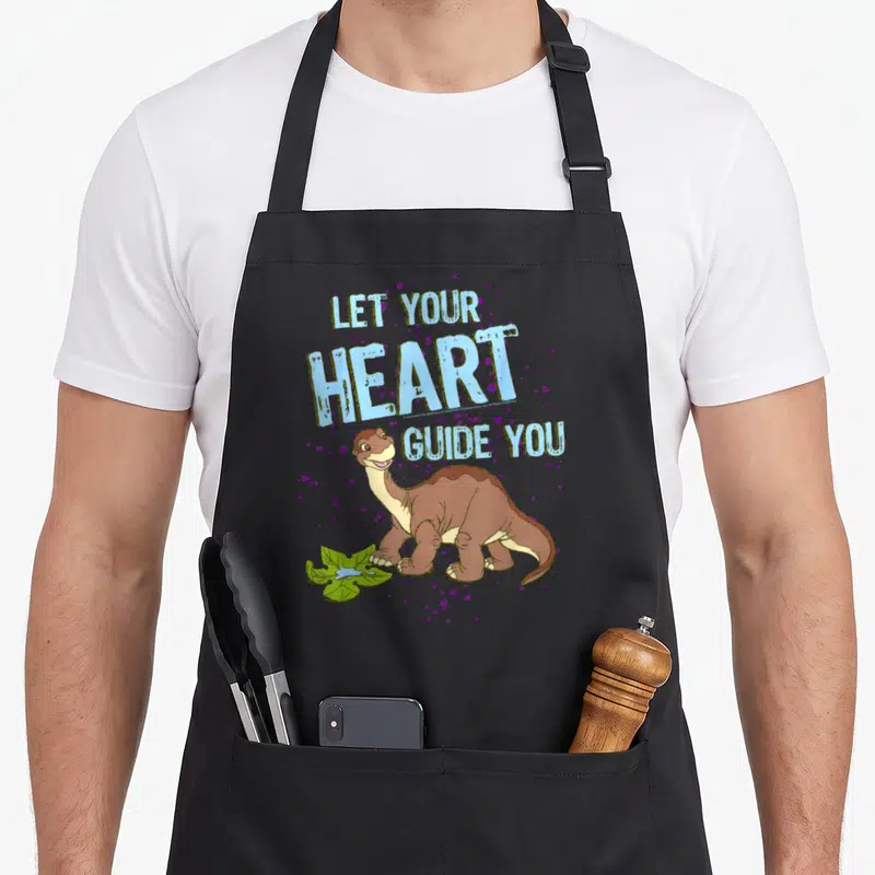 Land novelty apron - Cartoon brown dinosaur with LET YOUR HEART GUIDE YOU text and purple splatters — shown on a black cotton canvas apron against a white background