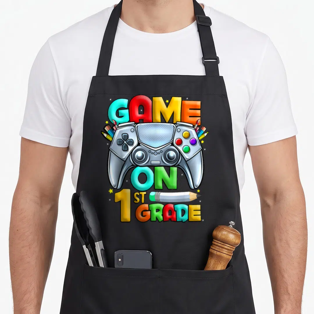 1st game aprons - Silver game controller with pencils and GAME ON 1ST GRADE text in multi-color cartoon style — shown on a black cotton canvas apron against a white background