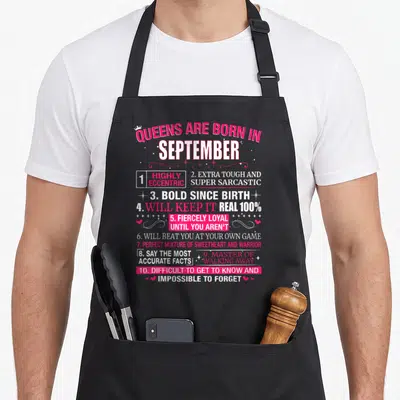 September queen apron - Pink and white text listing ten sarcastic traits scattered with hearts stars and a crown — shown on a black cotton canvas apron against a white background