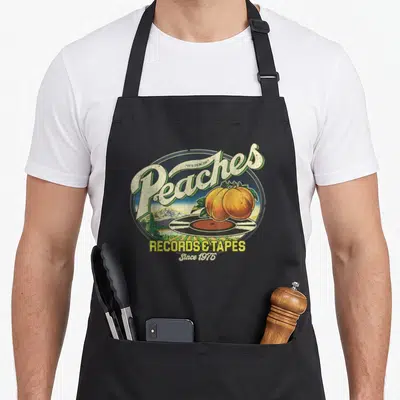 Peach record apron - Two peaches on vinyl record in retro oval with mountain background and text — shown on a black cotton canvas apron against a white background