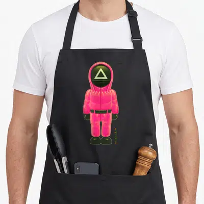 Soldier full body apron - Pink jumpsuit figure with triangle mask and vertical Korean text — shown on a black cotton canvas apron against a white background