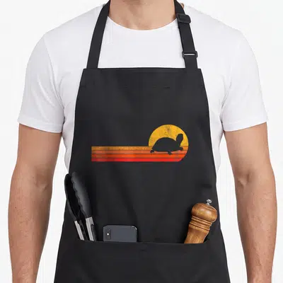 Cute turtle tortoise apron - Retro turtle silhouette against yellow sun and three orange red stripes — shown on a black cotton canvas apron against a white background
