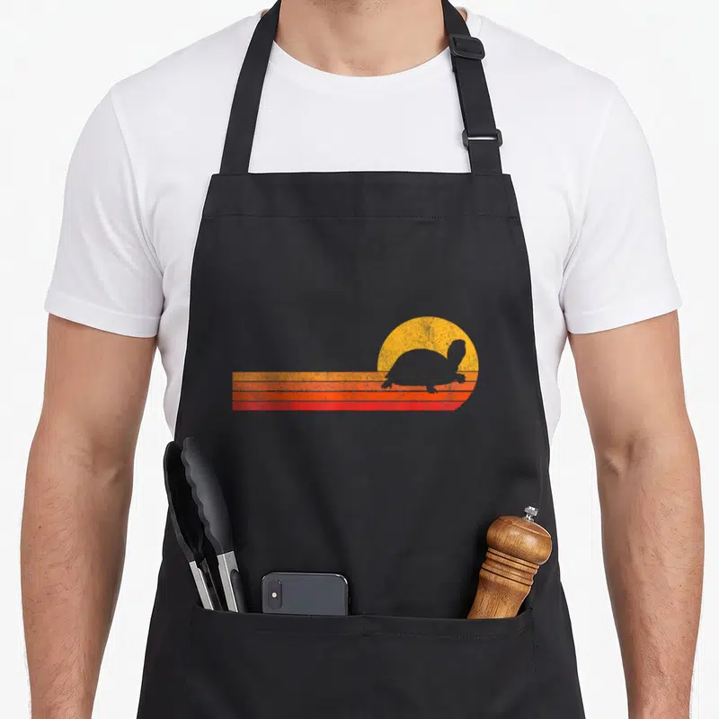 Cute turtle tortoise apron - Retro turtle silhouette against yellow sun and three orange red stripes — shown on a black cotton canvas apron against a white background