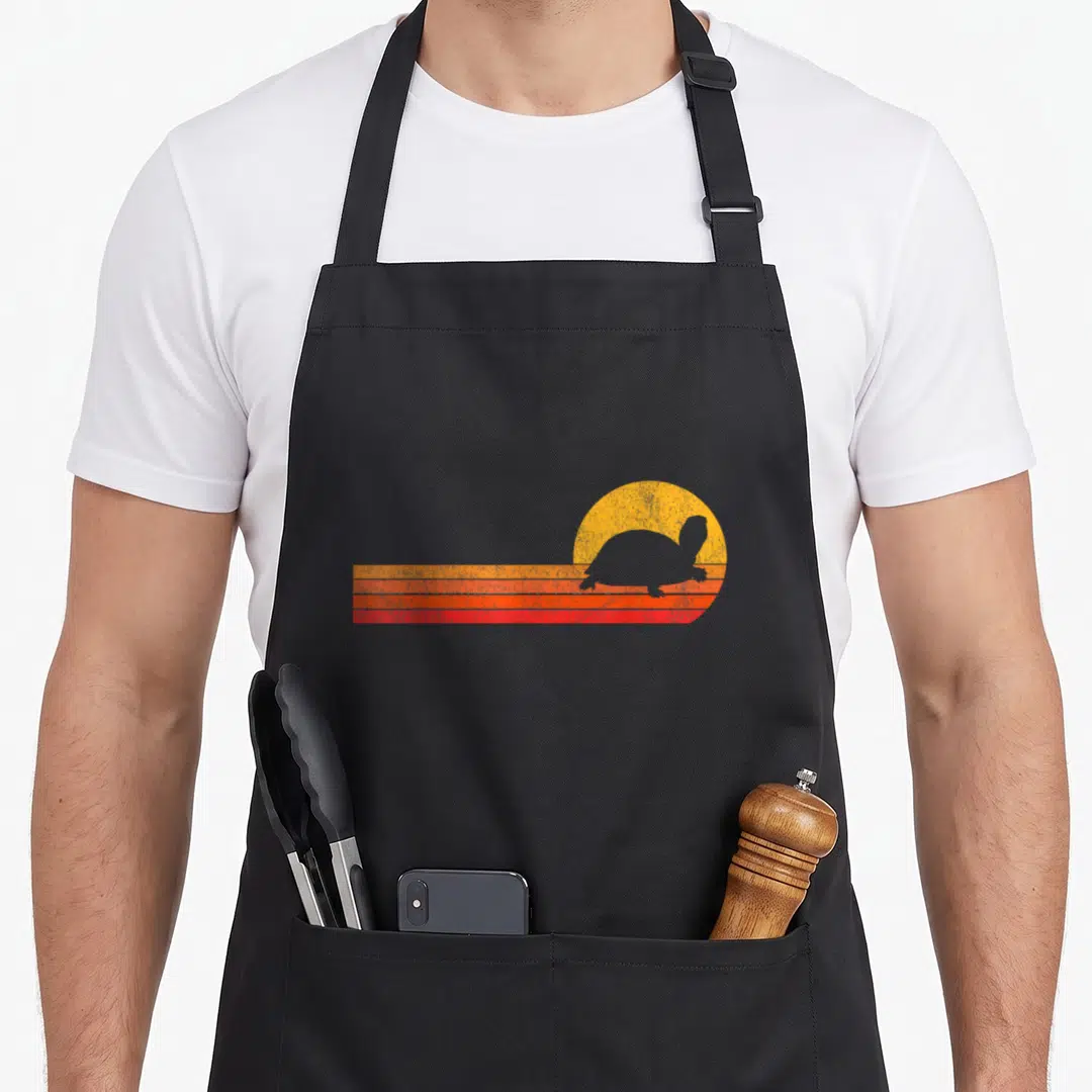 Cute turtle tortoise apron - Retro turtle silhouette against yellow sun and three orange red stripes — shown on a black cotton canvas apron against a white background