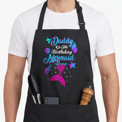 Birthday mermaid daddy apron - Pink mermaid tail with seashells and bubbles around stacked text — shown on a black cotton canvas apron against a white background