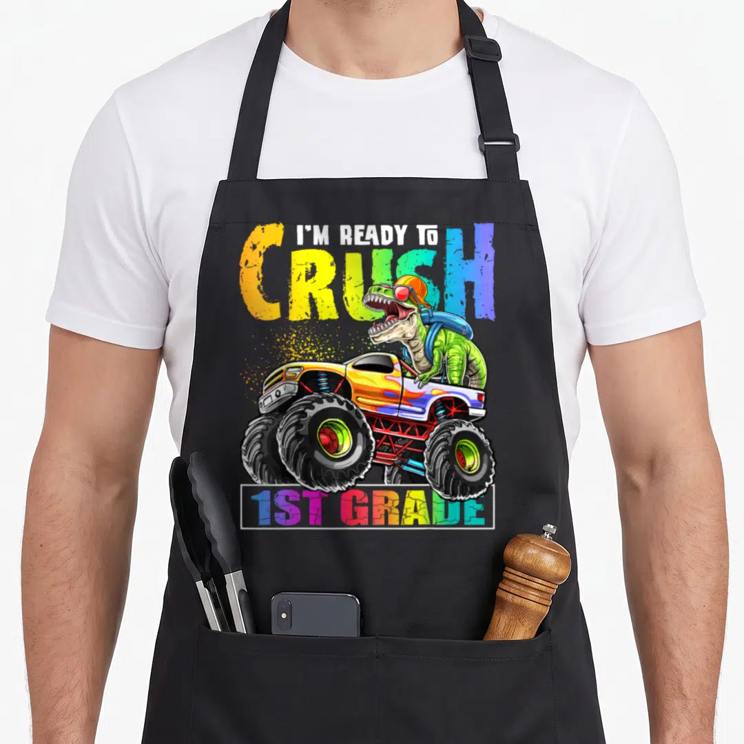 Crush 1st truck apron - Dinosaur in monster truck with I'M READY TO CRUSH 1ST GRADE text in rainbow colors — shown on a black cotton canvas apron against a white background