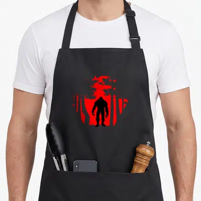 Scary wood apron - Black bigfoot silhouette standing in front of red circle and black tree shapes — shown on a black cotton canvas apron against a white background