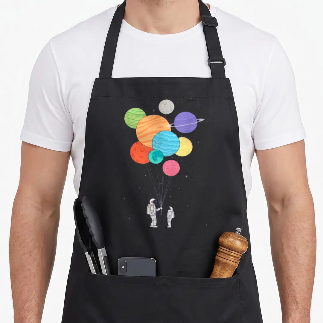 Funny planet balloon apron - Two astronauts holding strings attached to a cluster of colorful planets floating above — shown on a black cotton canvas apron against a white background