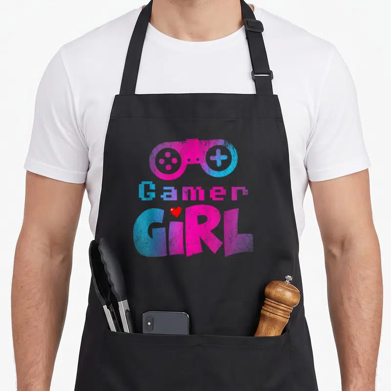 Game nerd apron - Pink blue game controller with pixel Gamer Girl text and red heart — shown on a black cotton canvas apron against a white background