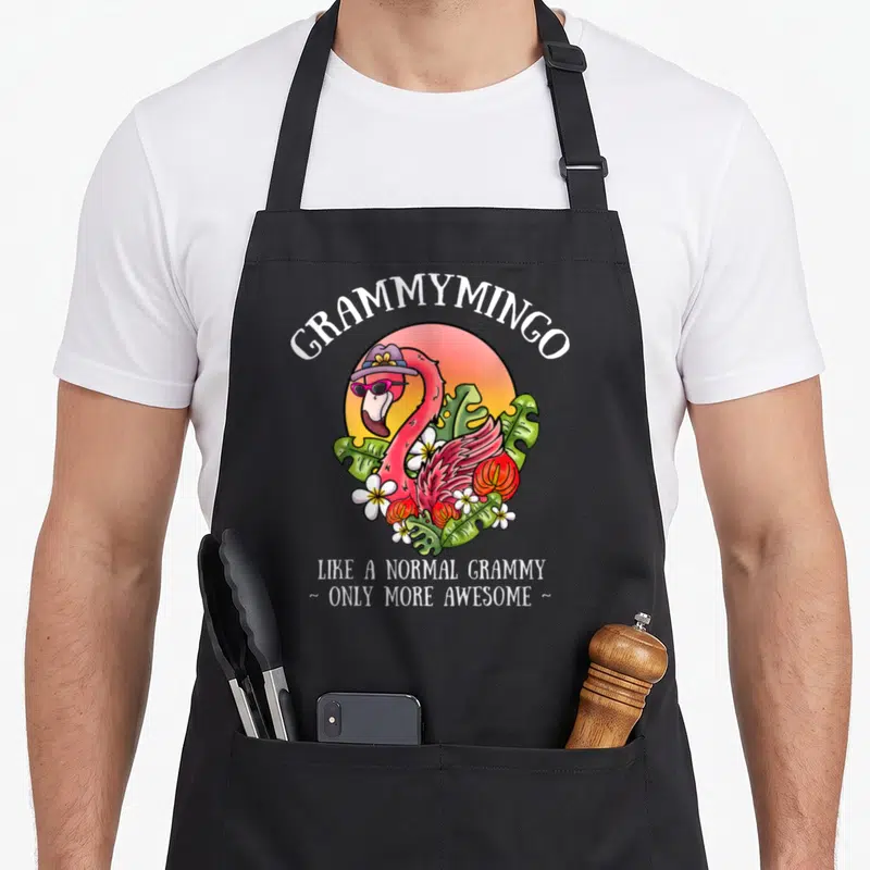 Grammymingo grandma apron - Pink flamingo wearing hat and sunglasses amidst green tropical leaves and white flowers — shown on a black cotton canvas apron against a white background