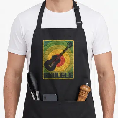 Uke pineapple apron - Grey ukulele silhouette over green yellow and red concentric circles with UKULELE text — shown on a black cotton canvas apron against a white background