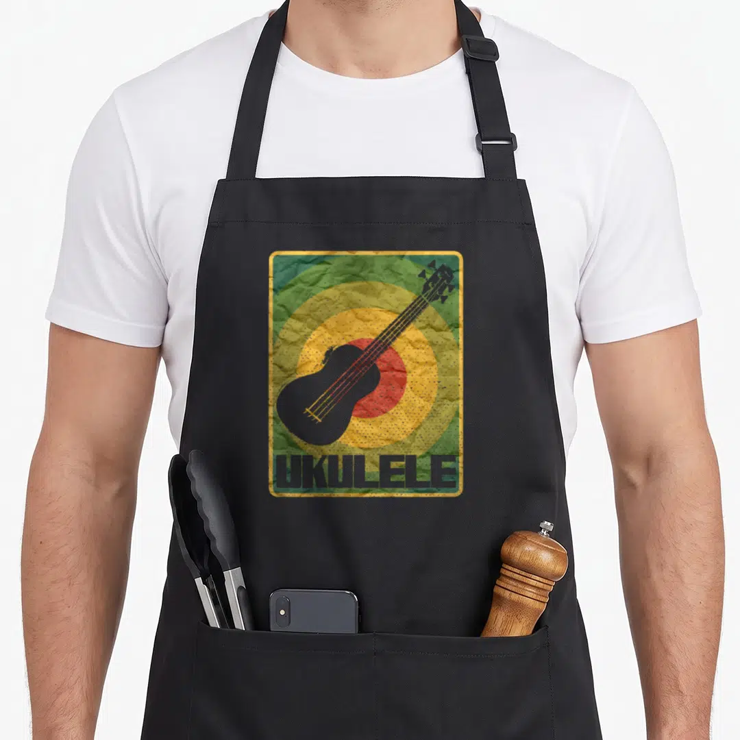 Uke pineapple apron - Grey ukulele silhouette over green yellow and red concentric circles with UKULELE text — shown on a black cotton canvas apron against a white background