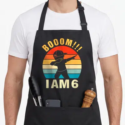 Dabbing chef outfit apron - Retro child silhouette dabbing in front of striped sun with BOOOM!!! I AM 6 text — shown on a black cotton canvas apron against a white background