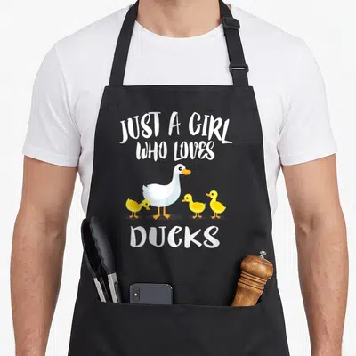 Funny owner duck apron - Cartoon white duck and three yellow ducklings under Just A Girl Who Loves Ducks text — shown on a black cotton canvas apron against a white background